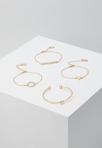 Quatre bracelets en or de designs variés, y compris un bracelet ouvert avec une pierre précieuse, des chaînes avec des accents circulaires et triangulaires, déposés sur une surface blanche.