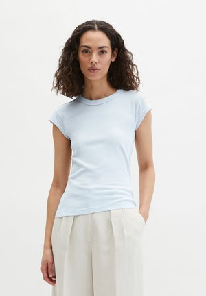 Kobieta o kręconych włosach, ubrana w dopasowaną, jasnoniebieską koszulkę z krótkim rękawem oraz beżowe, plisowane spodnie z wysokim stanem, z jedną ręką w kieszeni.