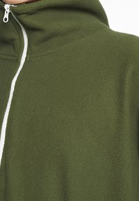 Olivgrön fleecehoodie med en texturerad yta, med en vit dragkedja och hög krage. Inga synliga mönster eller detaljer.