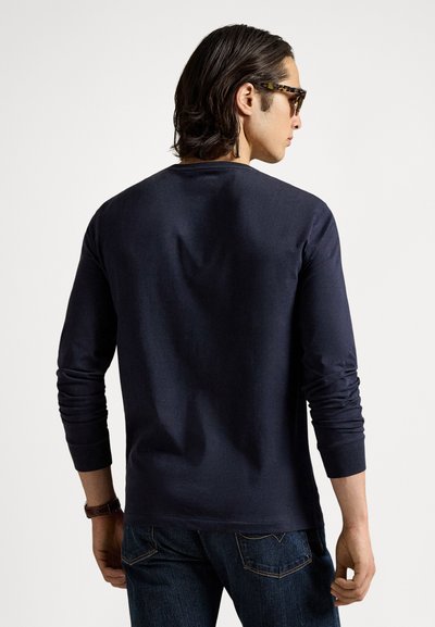 Tengerészkék hosszú ujjú póló puha anyagból, kerek nyakkivágással, testhez simuló mandzsettákkal és sima textúrával, sötét farmernadrággal párosítva.