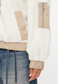Vrouwelijke fuzzy witte fleece jas met beige nylon accenten, comfortabele boorden en een zijzak met rits. Heeft een geribde zoom voor een aansluitende look.