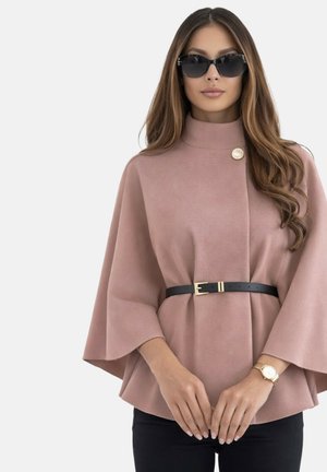 Femme aux longs cheveux portant des lunettes de soleil noires, un manteau cape mauve ceinturé avec des boutons dorés, un pantalon noir et une montre-bracelet en or.