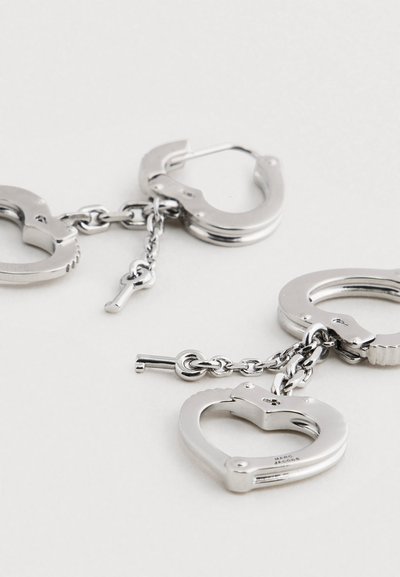 Strieborné kovové náušnice v tvare putiek a srdiečok, spojené reťazou, s malým detailom kľúča. Lesklý povrch, zložitý dizajn.