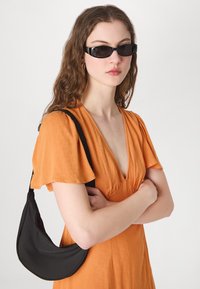 Schwarze rechteckige Sonnenbrille, oranges Kleid mit V-Ausschnitt und kurzen, schwingenden Ärmeln. Schwarze Schultertasche mit geschwungener Form.