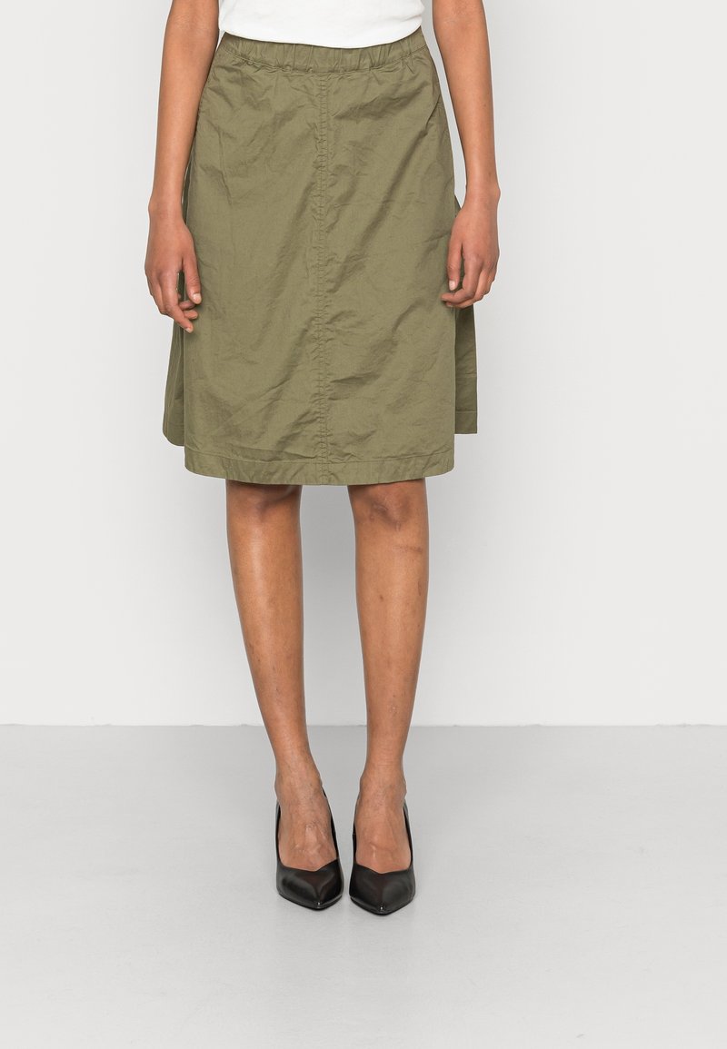 Femme portant une jupe longue jusqu'aux genoux de couleur vert olive avec une ceinture élastique et des talons noirs à bout pointu, debout devant un fond uni.