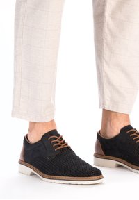 Schwarze perforierte Schuhe mit braunen Lederakzenten, runder Zehenform und beiger Sohle. Schnürsenkel in kontrastierendem Braun. Getragen mit hellen karierten Hosen.