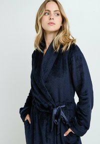 Peignoir en peluche bleu marine avec une finition texturée, col châle, taille ceinturée et poches latérales. Les manches longues et le tissu doux offrent confort.