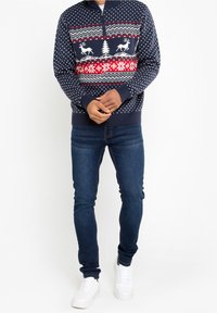 Marineblauer Strickpullover mit halbem Reißverschluss, versehen mit weißen Punkten und roten gemusterten Bändern, die Rentiere und Bäume darstellen; kombiniert mit schmal geschnittenen Jeans und weißen Sneakern.