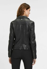 Zwarte leren jas met een getailleerd ontwerp, gestikte accenten op de schouders, ritsen aan de manchetten en een klassieke kraag. Gladde textuur met een lichte glans.