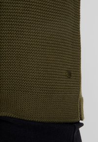 Olivgrüner, strukturiertes Strickgewebe über einem navyblauen Baumwollbereich, mit einem kleinen olivgrünen Akzentdetail nahe der Naht.