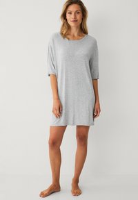 Vrouw die blootvoets staat in een losse, lichtgrijze, knielange jurk met halve mouwen tegen een effen witte achtergrond.