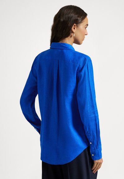 Μπλε πουκάμισο με μακριά μανίκια από υφασμάτινη υφή, με γιακά, μανσέτες με κουμπιά και πιέτα στην πλάτη. Στρογγυλεμένο τελείωμα.