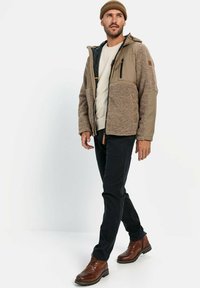 Tan- og grå fleecejakke med lynlås foran, to frontlommer og hætte, sammen med mørke bukser og brune støvler. Glat tekstur.