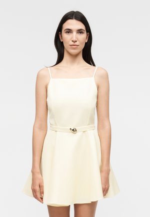 Žena s dugom tamnom kosom nosi haljinu bez rukava krem boje s tankim naramenicama i malim remenom s zlatnom kopčom u struku.