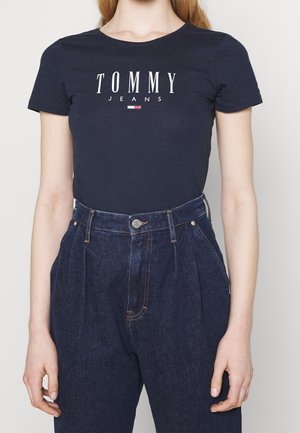 Femme portant un t-shirt Tommy Jeans bleu marine et un jean en denim bleu foncé plissé taille haute, debout avec les bras détendus.