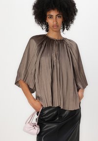 Gefaltete braune Bluse mit gerafftem Halsausschnitt, kurzen Ärmeln und leichtem Stoff; kombiniert mit einer kleinen rosa Handtasche und einem schwarzen Lederrock.