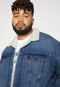 Mėlynas džinsinis švarkas su bežiniu pūkuotu apykakle, turintis mygtukų užsegimą, dvi priekinės kišenės ir prekės ženklo žymėje ant krūtinės.