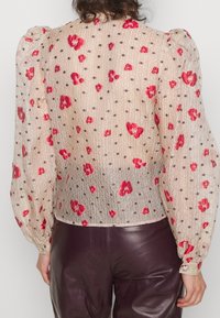 Durchsichtige Bluse mit Puffärmeln in Beige, verziert mit roten Blumenmustern und Punkten. Sie verfügt über einen strukturierten Stoff und dekorative Knopf-Details an den Manschetten.