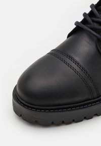 Zapato de cuero negro con un acabado suave, puntera redondeada y acentos cosidos. Presenta una suela de goma negra gruesa con un patrón de relieve.