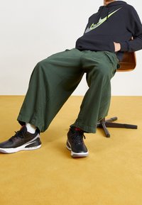 Pantalones holgados de color verde oscuro con un acabado texturizado, combinados con zapatillas negras con acentos rojos. Sudadera negra con un gran logo gris y neón.