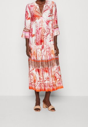 Kvinde iført en hvid midi-kjole med orange, pink og brune paisley- og dyreprintmønstre, kombineret med beige sandaler med blokhæl.