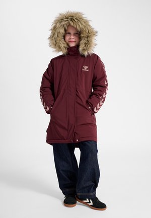 Barn iført en vinrød vinterjakke med pelsforet hætte, hænder i lommerne, mørke vidde jeans og sorte sneakers med hvide chevron-striber.