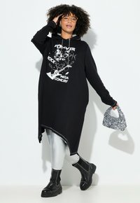 Zwarte oversized hoodie jurk met een grafische print, asymmetrische zoom, gecombineerd met glanzende zilveren leggings en chunky zwarte laarzen. Accessoires omvatten een pailletten tas.