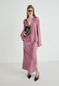 Donna in blusa mauve e gonna coordinata con dettagli in pizzo, tacchi a punta neri, occhiali da sole con tonalità blu, orecchini ad anello, mentre tiene una clutch verde e nera.