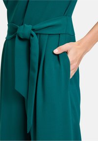 Teal jumpsuit met een strikdetail in de taille, gestructureerde stof, zijzakken en een soepele textuur. Het ontwerp omvat plooien bij de zak.