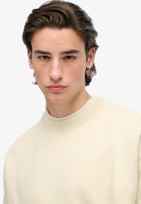 Giovane uomo con pelle chiara e capelli castani che indossa un maglione a maniche lunghe color crema con un piccolo logo ricamato sul petto.