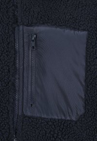 Tissu en polaire noire avec une poche rectangulaire zippée en nylon sur la poitrine et une fermeture éclair verticale sur le côté gauche.