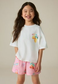 Lachend meisje met lang haar, dat een wit T-shirt met citroendruk en roze korte broek draagt, versierd met hartjes, kersen en zomerse motieven.