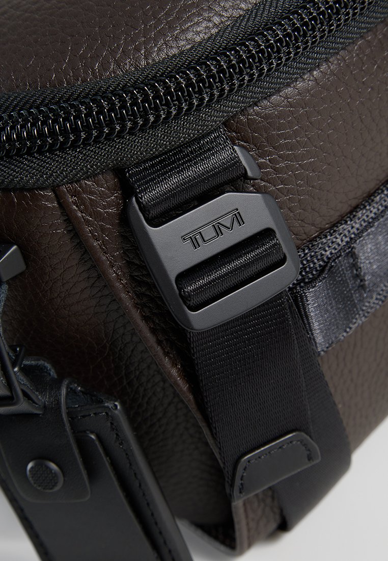 Braune Ledertasche mit schwarzen Reißverschlüssen, Gurtbandträger und TUMI-Logo-Beschlägen, die eine strukturierte Oberfläche und ein minimales Design präsentieren.