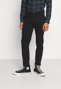 Svarta slim-fit byxor med en slät textur, i kombination med hög-top svarta canvas sneakers med en vit gummisula och logotyp.