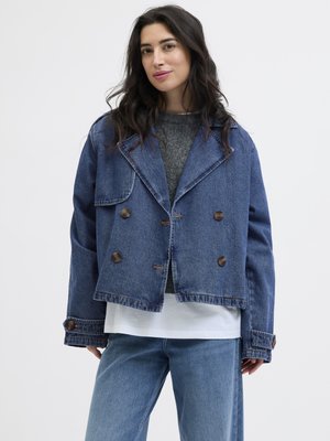Kvinne som har på seg en løs, dobbeltspent blå denimjakke over grå genser og hvit skjorte, kombinert med blå jeans, stående mot hvit bakgrunn.