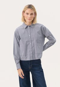 Szary i biały pasiasty shirt z kołnierzykiem, z przodu zapięcie na guziki oraz dwie duże kieszenie naszywane; ma długie rękawy i luźny krój.