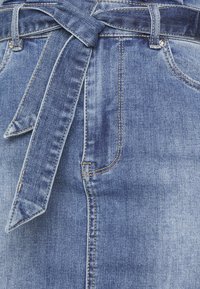 Spódnica dżinsowa w średnim odcieniu niebieskiego, z bocznymi kieszeniami i przewiązanym paskiem w talii. Gładka faktura z widocznymi detalami przeszycia.