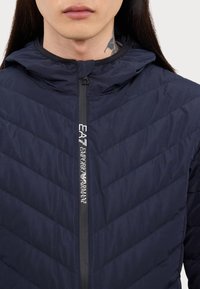 Ναυτικό μπλε μπουφάν puffer με κουκούλα, με φερμουάρ, σχέδιο σε διαγώνιες ραφές και λογότυπο στη ταινία του φερμουάρ.