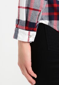 Röd, marinblå och vit rutigt flanellskjorta med vit manschett och knappdetalj, som visar en texturerad tyg och en åtsittande design.