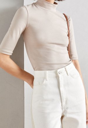 Osoba nosząca beżową bluzkę z golfem z rękawami do łokcia oraz białe spodnie z wysokim stanem, z brązowym paskiem przez ramię.