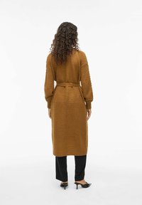 Cardigan en tricot marron avec un motif texturé, à manches longues et ceinture assortie, associé à un pantalon noir et des talons.
