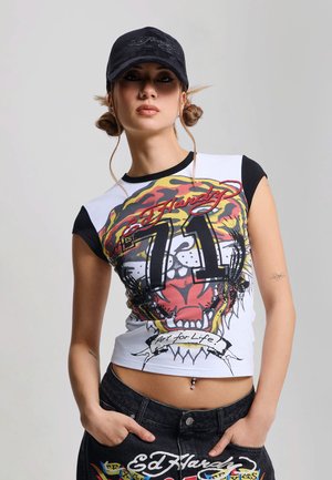 Mujer joven con moños laterales que lleva gorra negra, camiseta blanca Ed Hardy con gráfico de tigre, vaqueros negros y piercing en el ombligo, posando contra un fondo gris.