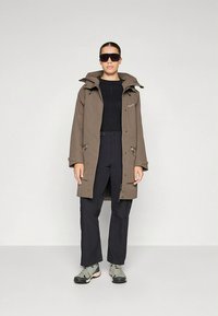 Giacca marrone impermeabile con cappuccio, tasche laterali con zip, indossata sopra una maglia nera e pantaloni abbinati, abbinata a scarpe da trekking grigie.