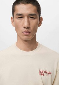 Cream-colored cotton t-shirt with a crew neckline, featuring red text graphic "EQUESTRIAN PRIDE" on the left chest area.