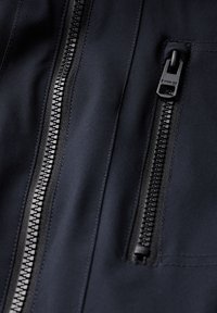 Veste marine en tissu brillant, avec col montant, deux poches zippées avec quincaillerie noire, et coutures texturées le long des bords.