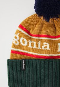 Gorro de malha da Patagonia com riscas amarelo mostarda, vermelho e branco, punho canelado verde escuro e pompom azul marinho no topo.