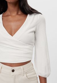 White, textured wrap top with a deep V-neck and three-quarter puff sleeves, paired with light beige high-waisted pants.