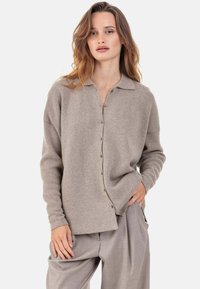 Cardigan en maille beige avec col, boutonnage sur le devant et coupe décontractée. Texture douce, manches longues, porté avec un pantalon beige plissé.