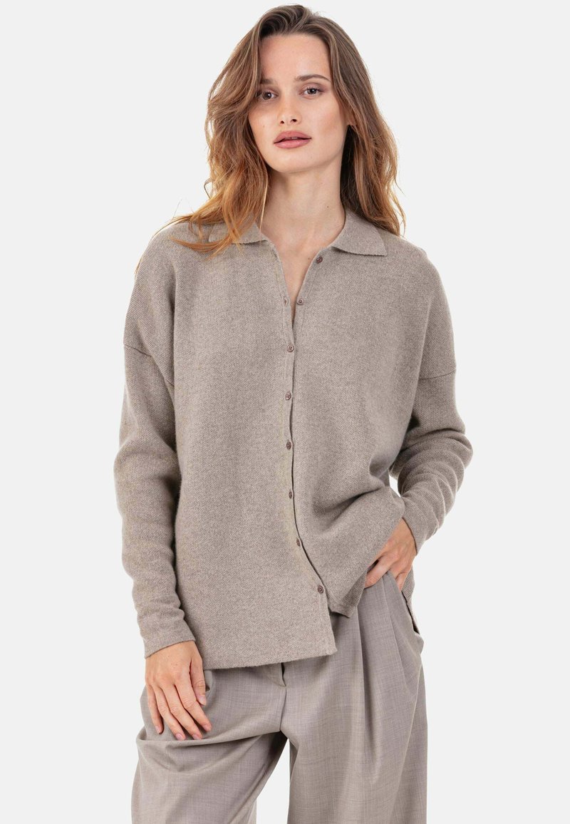 Cardigan en maille beige avec col, boutonnage sur le devant et coupe décontractée. Texture douce, manches longues, porté avec un pantalon beige plissé.