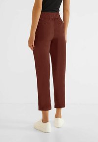 Person klädd i högmidjade, korta bruna byxor med elastisk midja, svart topp och vita sneakers, står på ett vitt golv mot en vit bakgrund.